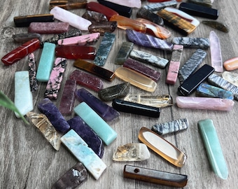 May include: A collection of rectangular gemstone pendants in various colors, including pink, purple, green, and black. The pendants are arranged on a wooden surface, showcasing their diverse textures and hues. Some pendants have a hole drilled through them.