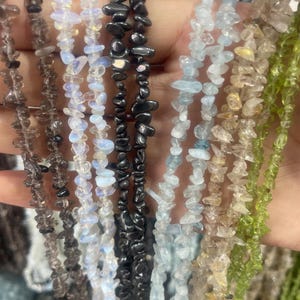 Op de afbeelding: Meerdere strengen van onregelmatige, getrommelde edelsteensplinters in diverse kleuren, waaronder rookbruin, iriserend wit, metallic zwart, lichtblauw, doorschijnend beige en levendig groen, geregen aan dunne, flexibele sieradendraad.