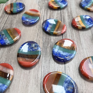 May include: Seven chakra stones, each with a different colour and pattern, arranged on a wooden surface. The stones are oval-shaped and polished.