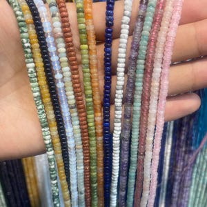 Puede incluir: Una colección de múltiples hilos de pequeñas cuentas de piedras preciosas, redondas y lisas, en una variedad de colores que incluyen verde, amarillo, negro, azul, naranja, blanco, morado y rosa, dispuestas verticalmente en abanico.