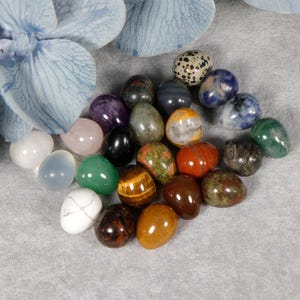 Op de afbeelding: Een verzameling gepolijste edelsteen eieren in verschillende kleuren, waaronder amethist, tijgeroog en groene aventurijn. De eieren zijn gerangschikt op een grijze ondergrond, met een blauw bloemenaccent op de achtergrond. Elk ei heeft een uniek patroon en textuur.