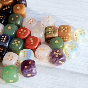 May include: A collection of gemstone dice in various colors, including green, red, pink, and purple. The dice have gold-colored dots and are scattered on a light-colored surface. The dice are a mix of opaque and translucent stones.
