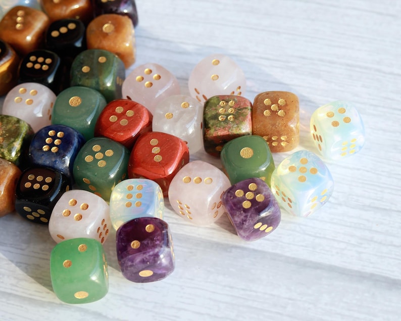 May include: A collection of gemstone dice in various colors, including green, red, pink, and purple. The dice have gold-colored dots and are scattered on a light-colored surface. The dice are a mix of opaque and translucent stones.