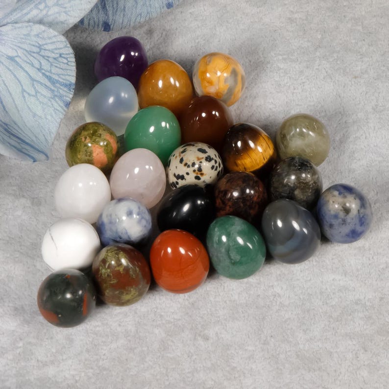 Puede incluir: Una colecci&oacute;n de huevos de piedras preciosas pulidas en varios colores, incluyendo p&uacute;rpura, verde, naranja y patrones moteados. Los huevos est&aacute;n dispuestos sobre una superficie gris claro, mostrando sus diversas texturas y tonalidades. Las piedras preciosas parecen lisas y reflectantes.