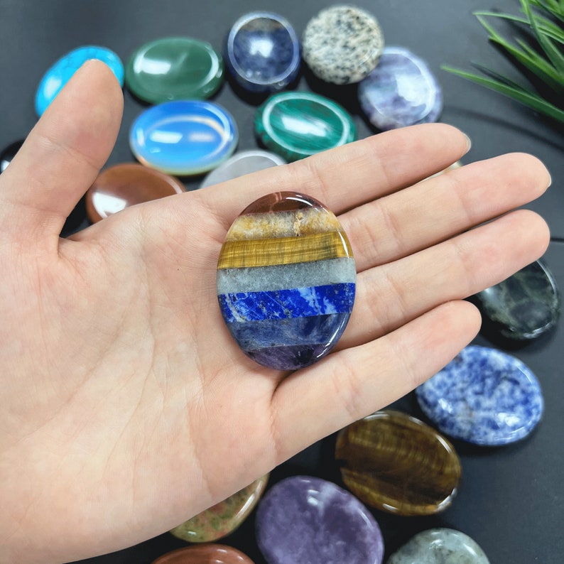 K&ouml;nnte beinhalten: Ein sieben-Chakra-Handstein mit Schichten aus rotem Jaspis, Karneol, gelbem Tigerauge, klarem Quarz, Lapislazuli, Amethyst und klarem Quarz. Der Stein ist oval geformt und wird in einer Hand gehalten.