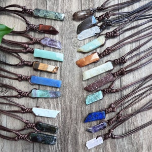Puede incluir: Una colección de 18 colgantes de piedras preciosas en cordones de cuero marrón. Los colgantes son de diferentes formas y tamaños, y están hechos de varios tipos de piedras preciosas, incluyendo amatista, ágata y turquesa.