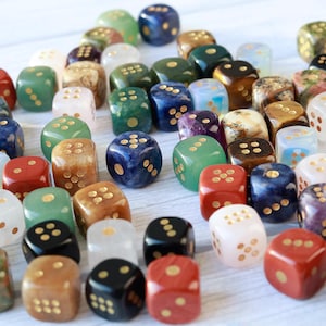 May include: A collection of gemstone dice in various colors, including red, green, blue, and white. Each die has gold-colored dots. The dice are scattered on a light-colored surface, creating a visually appealing arrangement for tabletop games.
