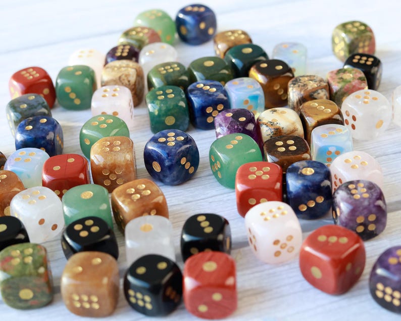 May include: A collection of gemstone dice in various colors, including red, green, blue, and white. Each die has gold-colored dots. The dice are scattered on a light-colored surface, creating a visually appealing arrangement for tabletop games.
