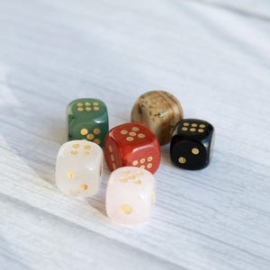 May include: Six colorful dice in various colors, including green, red, black, white, and a speckled brown. Each die has gold-colored dots. The dice are arranged on a light-colored wooden surface.