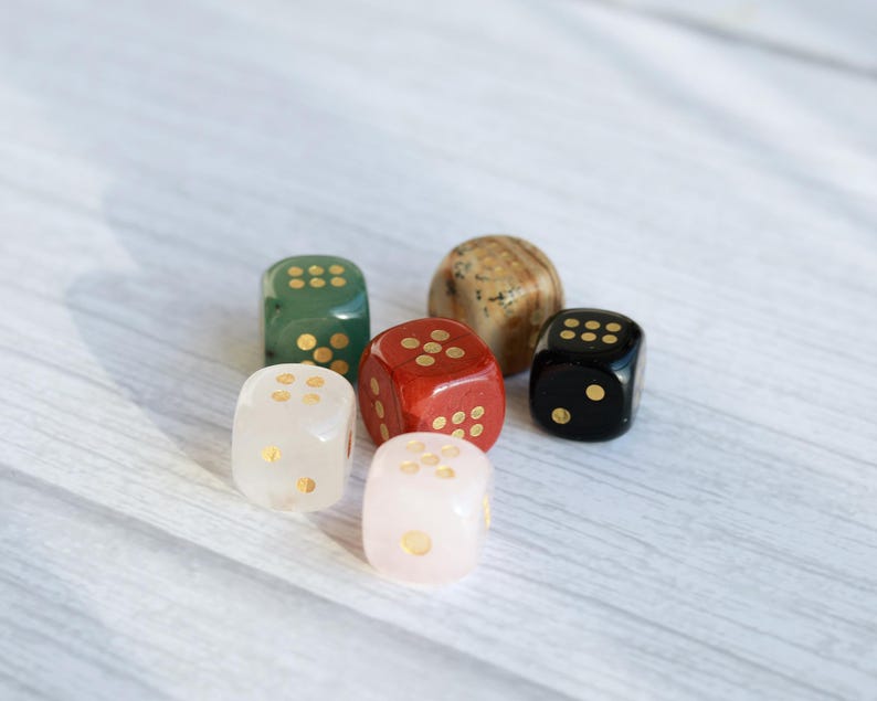 May include: Six colorful dice in various colors, including green, red, black, white, and a speckled brown. Each die has gold-colored dots. The dice are arranged on a light-colored wooden surface.