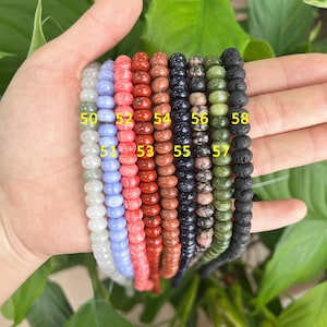 May include: A close-up of ten strands of beads, each with a different color and texture. The beads are numbered 50 through 58. The beads are on a hand, and the background is a green plant.