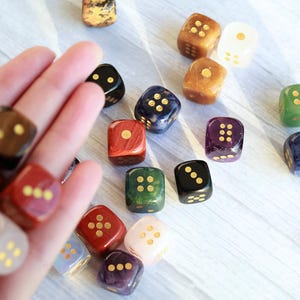 May include: A collection of colorful dice with gold-colored pips. The dice are in various colors, including red, green, black, and purple. Some dice are held in a hand, while others are scattered on a light-colored surface.