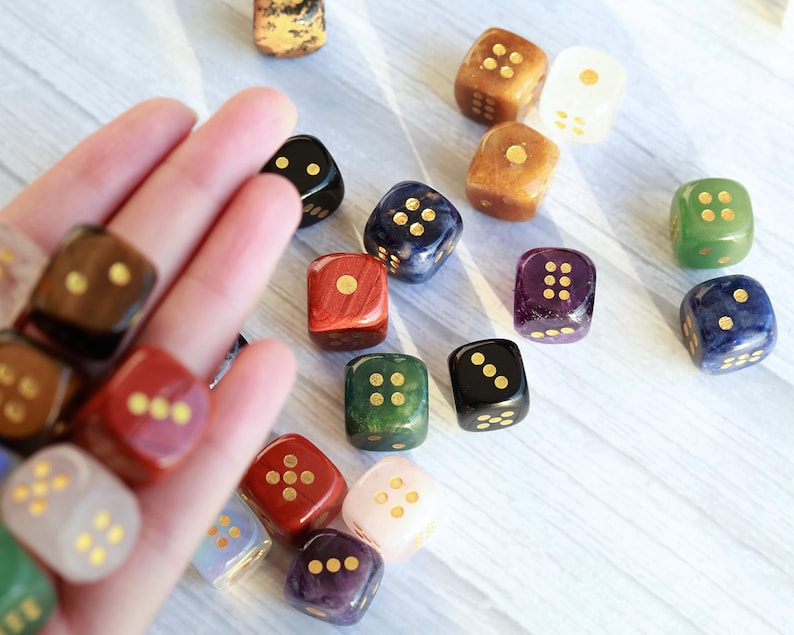 May include: A collection of colorful dice with gold-colored pips. The dice are in various colors, including red, green, black, and purple. Some dice are held in a hand, while others are scattered on a light-colored surface.
