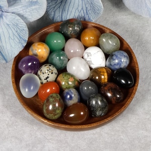 Puede incluir: Un cuenco de madera lleno de varios huevos de piedras preciosas pulidas en tonos verdes, morados, naranjas, blancos y marrones. Los huevos est&aacute;n dispuestos en un patr&oacute;n circular, mostrando sus colores y texturas &uacute;nicos. El cuenco se encuentra sobre una superficie gris claro.