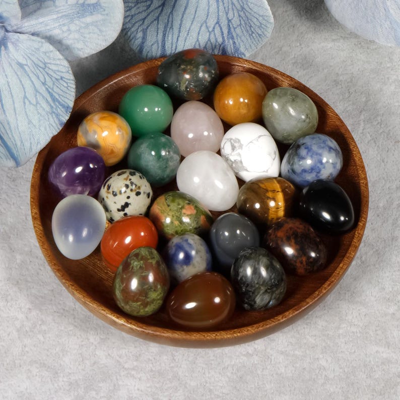 Puede incluir: Un cuenco de madera lleno de varios huevos de piedras preciosas pulidas en tonos verdes, morados, naranjas, blancos y marrones. Los huevos est&aacute;n dispuestos en un patr&oacute;n circular, mostrando sus colores y texturas &uacute;nicos. El cuenco se encuentra sobre una superficie gris claro.