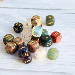 May include: A collection of various gemstone dice in different colors, including green, red, blue, and brown. Each die has gold-colored pips. The dice are scattered on a white wooden surface.