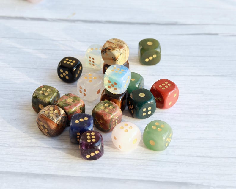 May include: A collection of various gemstone dice in different colors, including green, red, blue, and brown. Each die has gold-colored pips. The dice are scattered on a white wooden surface.