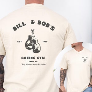 May include: Cream-colored t-shirt featuring a boxing gym graphic. The design includes the text "BILL & BOB'S" above a pair of boxing gloves, with "EST 1935" below. The words "BOXING GYM" and "AKRON, OH" are also printed on the shirt.