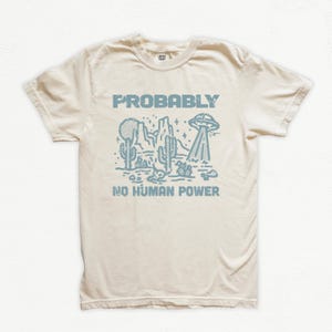Pode incluir: Camiseta creme com um gráfico azul claro. O gráfico apresenta uma paisagem desértica com cactos, montanhas, um OVNI e o texto "PROBABLY NO HUMAN POWER."