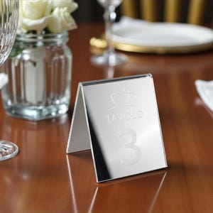 Könnte beinhalten: Ein silberner Tischaufsteller mit dem Wort "Tavolo" und der Zahl "3" eingraviert. Der Tischaufsteller ist im Stil einer Zeltkarte gehalten und steht auf einem Holztisch. Im Hintergrund befindet sich eine Glasvase mit weißen Blumen.