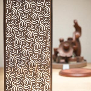 Op de afbeelding: Een rechthoekig houten paneel met een herhalend patroon van wervelende, onderling verbonden lijnen. De lijnen zijn lichtbruin en de achtergrond is wit. Het paneel is omlijst in donkerder bruin hout. Op de achtergrond is een vage houten sculptuur te zien.