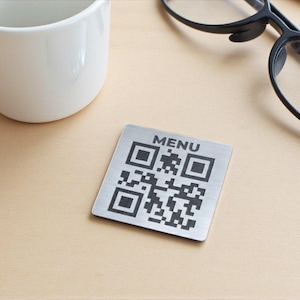 Può includere: Targhetta quadrata in metallo spazzolato con codice QR e la scritta «MENU» in alto. La targhetta è su una superficie chiara, accanto a una tazza bianca e un paio di occhiali neri.