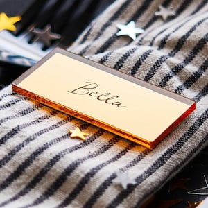 Könnte beinhalten: Goldene Platzkarte mit schwarzem Rand und dem Namen "Bella" in Kursivschrift.
