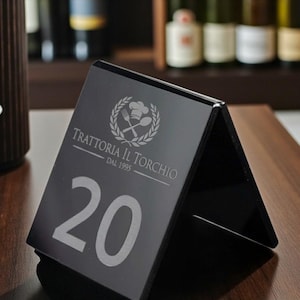 Könnte beinhalten: Eine schwarze, dreieckige Tischnummer mit der Zahl 20 in Weiß. Das Schild trägt den Text "Trattoria Il Torchio Dal 1995" und ein Logo mit Kochmütze, Gabel und Löffel. Das Schild steht auf einem Holztisch.