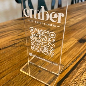 Può includere: Un cartello in acrilico trasparente con il testo "ember - SIP | SWIM | SUNSETS -" e un codice QR. Il testo "@CambriaMadeiraHotel" è sotto il codice QR.