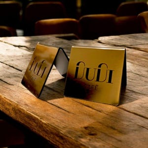 Op de afbeelding: Twee gouden metalen tafelschilderingen met de tekst "DUDI CAFE" op een houten tafel.