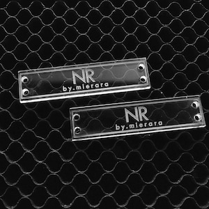 Könnte beinhalten: Zwei rechteckige, transparente Acrylschilder mit abgerundeten Ecken. Jedes Schild hat zwei Löcher zum Befestigen. Der Text "NR by.mierara" ist auf jedem Schild graviert.