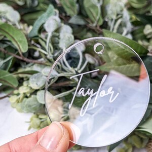 Könnte beinhalten: Transparenter runder Acryl-Anhänger mit dem Namen "Taylor" in Schreibschrift. Der Anhänger hat ein kleines Loch zum Aufhängen. Der Hintergrund zeigt grünes Laub.