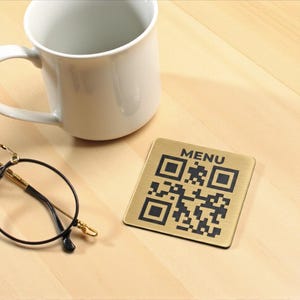Può includere: Un sottobicchiere quadrato color oro con un codice QR e la parola "MENU" in nero. Una tazza bianca e occhiali con montatura nera e lenti rotonde sono anche su una superficie di legno chiaro.