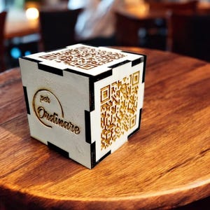 Può includere: Un cubo di legno bianco con un codice QR nero e oro e il testo "per Ordinare" su un lato.