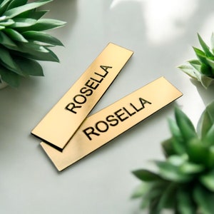 Könnte beinhalten: Zwei rechteckige Namensschilder aus goldenem Metall mit dem Namen "Rosellla" in schwarzen Lettern graviert.