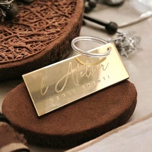 Op de afbeelding: Een gouden metalen sleutelhanger met een rechthoekig label waarop "L'Atelier Degli Eventi" staat. De sleutelhanger ligt op een bruine suède ondergrond.