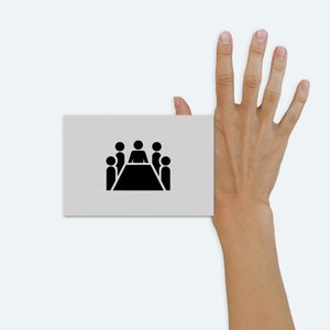 Può includere: Un cartoncino bianco con un'icona nera di un tavolo con cinque persone sedute intorno. Il cartoncino è tenuto da una mano con cinque dita estese.
