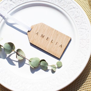 May include: A wooden place card with the name Amelia engraved on it. The card is tied with a white ribbon and is sitting on a white plate.