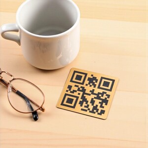 Può includere: Una tazza di ceramica bianca, un sottobicchiere quadrato color oro con un codice QR nero e un paio di occhiali con montatura dorata poggiano su una superficie di legno chiaro. Il sottobicchiere è quadrato e presenta un complesso motivo di quadrati neri e oro.