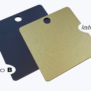 Può includere: Due etichette metalliche quadrate con un foro nella parte superiore. Un'etichetta è blu scuro e l'altra è dorata. L'etichetta dorata ha il testo "lato A" sul lato destro e l'etichetta blu ha il testo "lato B" sul lato sinistro.