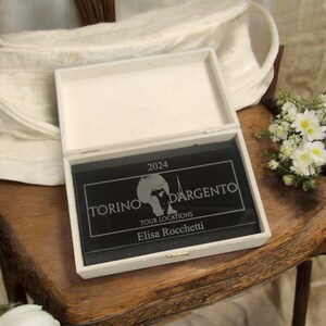 Puede incluir: Una caja de madera blanca con una placa de acrílico negro en su interior. La placa tiene una inscripción plateada que dice "2024 TORINO D'ARGENTO TOUR LOCATIONS Elisa Rocchetti".