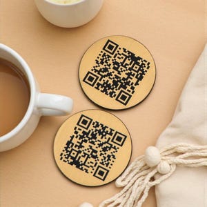 Può includere: Due sottobicchieri rotondi in legno chiaro con motivi a codice QR neri. I sottobicchieri sono su una superficie beige, vicino a una tazza bianca con una bevanda marrone. Sono visibili anche una piccola ciotola bianca e un tovagliolo color crema.