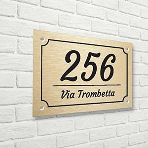 May include: A gold-colored metal house number sign with black lettering. The number 256 is displayed in a cursive font. The text "Via Trombetta" is displayed below the number.