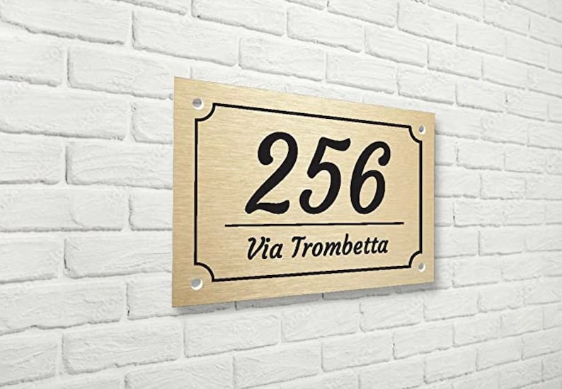 May include: A gold-colored metal house number sign with black lettering. The number 256 is displayed in a cursive font. The text "Via Trombetta" is displayed below the number.