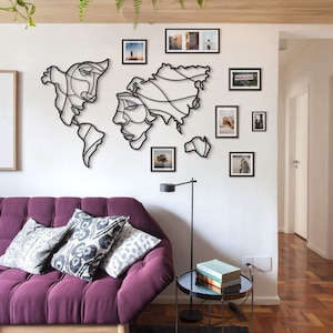 Op de afbeelding: Een zwarte metalen wandkunst sculptuur van een wereldkaart met twee gezichten, omringd door ingelijste foto's in een woonkamer met een paarse bank en een zwarte bijzettafel.