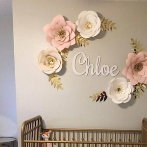 Può includere: Un arredamento per pareti di una nursery con una ghirlanda di fiori di carta con fiori rosa e bianchi e foglie dorate. Il nome "Chloe" è scritto in lettere bianche sul muro.