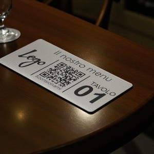 Può includere: Un segnaposto rettangolare in metallo spazzolato con la parola "Logo" in corsivo, un codice QR e il testo "Il nostro menu TAVOLO 01". Il segnaposto è su un tavolo di legno scuro, con calici sullo sfondo.