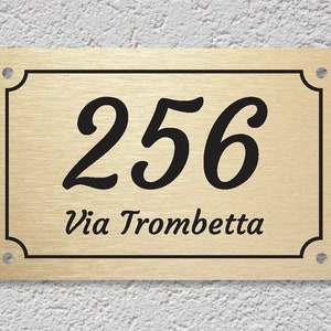 May include: Gold metal house number sign with black lettering. The number 256 is displayed in a cursive font. The text "Via Trombetta" is displayed below the number.