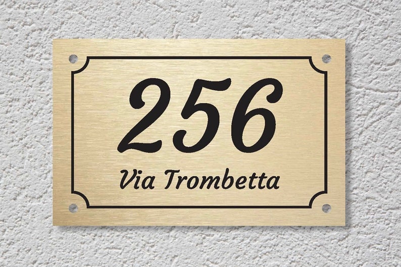 May include: Gold metal house number sign with black lettering. The number 256 is displayed in a cursive font. The text "Via Trombetta" is displayed below the number.