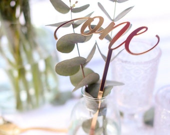 Peut inclure: Numéro de table 'un' en or avec une tige dorée, placé dans un vase en verre transparent avec de la verdure d'eucalyptus.